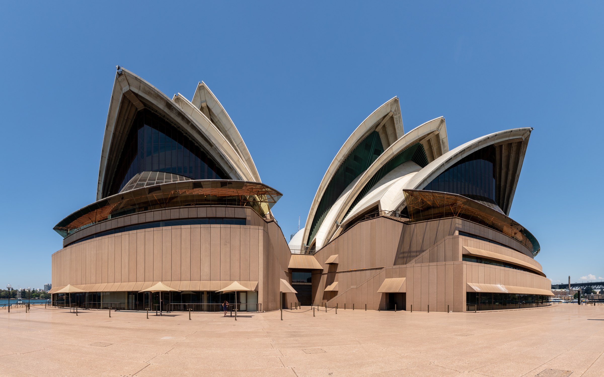 The white shell roofs and glass walls of Sydney Opera House, with people walking across the forecourt.